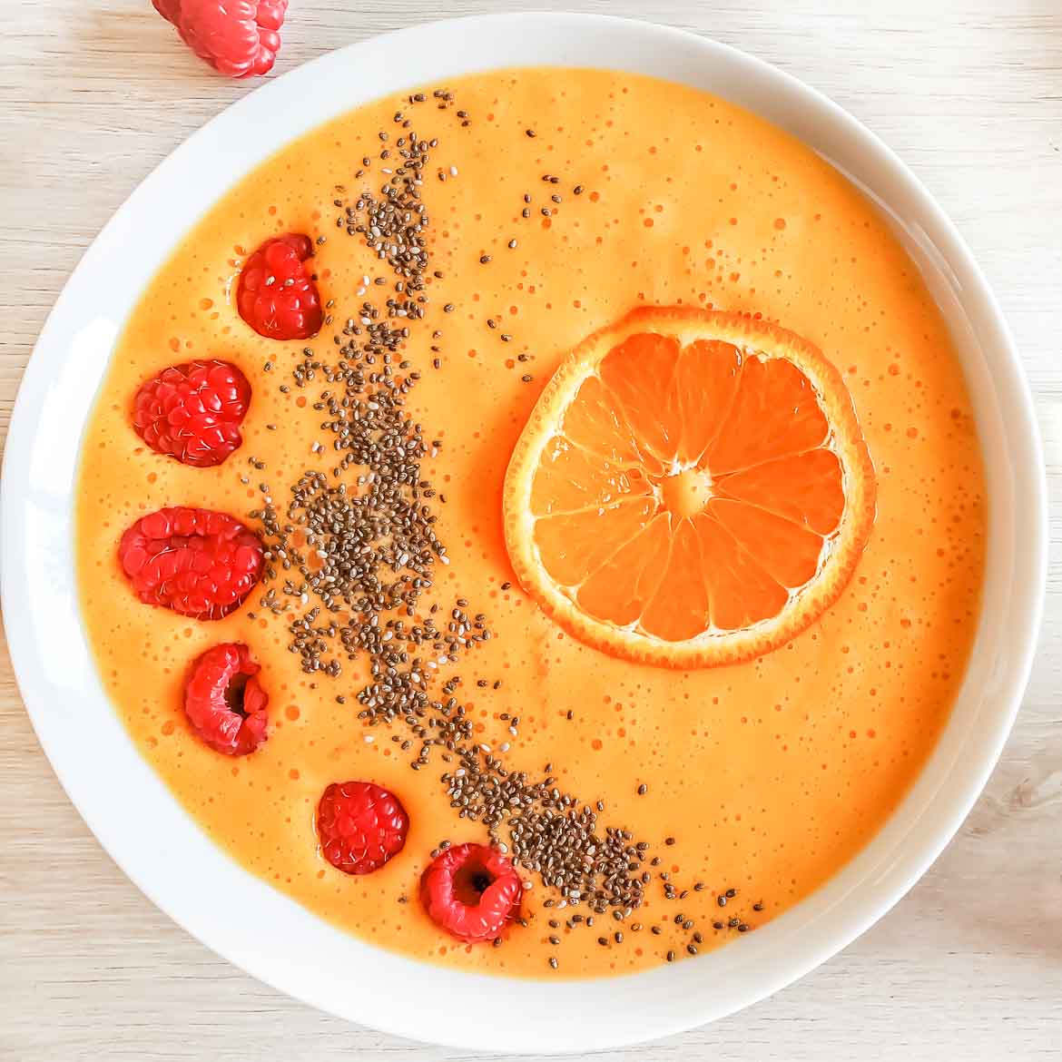 mango smoothie bowl with banana and orange, garnished with orange slice, raspberry, and chia seeds